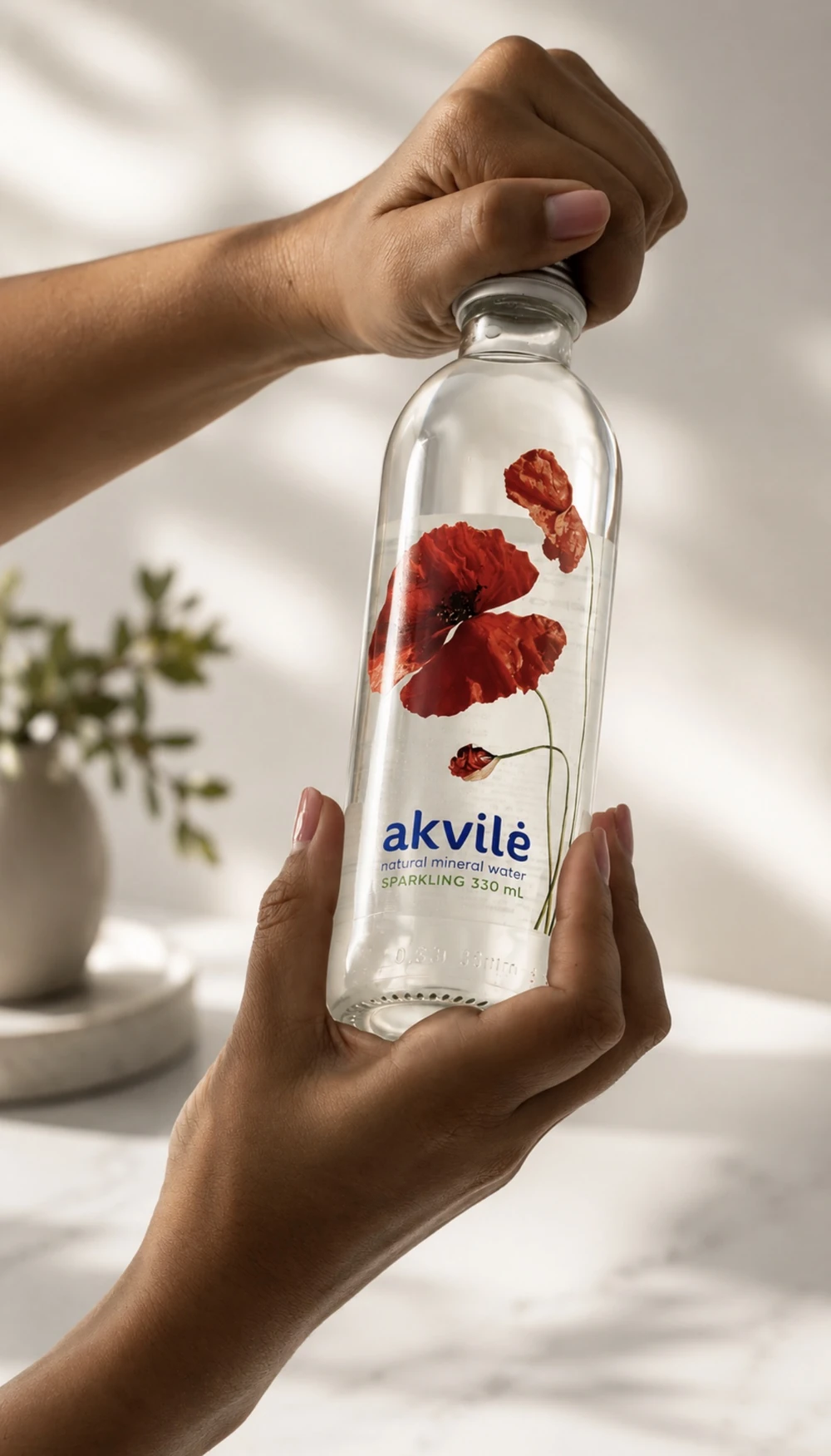 Two hands opening an akvilė sparkling 330 mL glass bottle in soft daylight